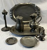 Full view of all 8 antique pewter pieces including 2 oil lamps, 2 large trays, 2 pitchers on a tray, and 2 small round trays.