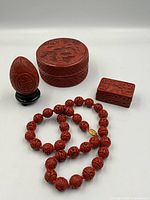 Full lot of four cinnabar red lacquer Asian decorative items: one round box, one rectangular box, one egg with stand, and one beaded necklace coiled in front