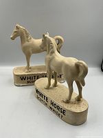 Pair of ceramic horse advertising figures labeled White Horse Scotch Whisky, cream colored with wear and paint chipping.