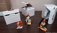 Three Disney ornaments displayed on table with their original open boxes behind: Pocahontas sitting, Lady dog in front with snowman charm, Pinocchio standing holding a scroll.