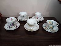 Photo showing all five fine bone china cups and matching saucers with floral patterns and gold trim on a dark wood surface.