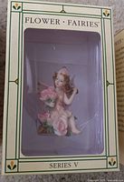 Figurine visible inside a box with a window. Fairy holding pink flowers, seated, with floral details, pastel colors, and wings.