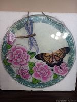 Front view of the round glass window ornament showing the butterfly and dragonfly design among pink roses and a decorative border with chain attached