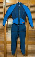 Full front view of a ladies wetsuit hanging on a closet door showing blue arms and upper chest with black torso and legs.