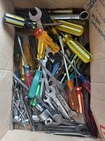 Top-down view of a cardboard box filled with assorted old hand tools including screwdrivers with yellow, amber, red, black, and green handles, wrenches, drill bits, and sockets.
