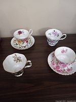 Four bone china cups and two saucers displaying various floral patterns and gold trims on a wooden surface.