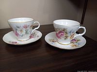 Two Sadler Wellington fine bone china cups and saucers, each with unique floral designs and gold rim decoration, displayed on a wooden surface.