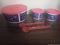 Three Tupperware bowls of varying sizes with locking pink lids that feature a purple floral design on the bowl sides, accompanied by a pink plastic salad spoon and fork.