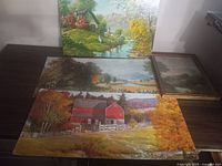 Four vintage cardboard prints arranged on a dark table showing rural landscapes in color, including a red barn and river with trees.