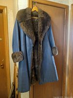 Full view of the vintage blue leather coat with brown rabbit fur collar and cuffs hanging on a wooden hanger on a door.