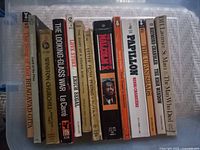 Side view of a row of vintage paperback books with visible titles including Bhagavad-Gita, The Looking Glass War, Malcolm X, Papillon, and The Upanishads.