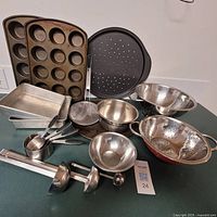 Image shows a collection of metal baking and kitchen tools including cake pans, muffin tray, mixing bowls, strainers, ladles, measuring cup, and cooling rack all arranged on a surface.