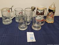 Seven assorted vintage beer mugs displayed on a blue cloth, including glass mugs and ceramic steins with pewter lids and decorative scenes.