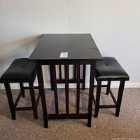 Front view of dark brown wood bistro table with two black padded stools tucked underneath, showing overall set appearance and size proportion.