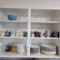 Shelves with dishes including clear wine glasses on the top shelf, assorted mugs and cups on the middle shelf, white bowls and drinking glasses below, and stacks of white plates to the right