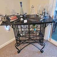 Front view of the wrought-iron wine rack cabinet with bar accessories displayed on black marble top, including glassware, decanter, cocktail shaker, and other bar tools.