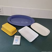 Four ceramic items on counter including large blue oval Emile Henry serving platter, yellow Emile Henry butter dish with lid, and two medium rectangular dishes, one green and white, one white.