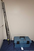 Photo of two fishing rods standing upright next to a closed blue tackle box on the floor against a white wall.