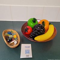 Photo showing red glass fruit bowl with green apple, orange, banana, black grapes, red pomegranate and olive wood bowl with various colorful glass candy figurines.