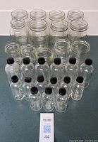 Photo of 15 clear glass mason jars and 13 clear glass medicine jars with black caps arranged in rows on a surface.