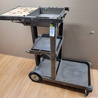 Side angle view of the three-tier gray plastic janitorial cart on a wooden floor, showing top shelf with holes and middle shelf.