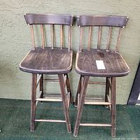 Front view of two wooden bar stools with vertical spindle backrests and flat seats against a green wall and green carpeted floor.