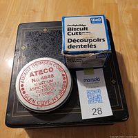 Tin of Ateco No. 4848 Medium Aspic Cutters and box of Wilton Biscuit Cutters placed on decorative black tin box on wooden floor.