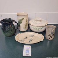 Front view of 5 assorted pottery pieces: green-blue pitcher, tall fern vase, covered bowl, small floral pitcher, and oval platter.