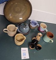 Overhead view of 11 various pottery items on tabletop showing large brown bowl, several pitchers, mugs, a lidded pot and small vessels grouped together with MaxSold tag.