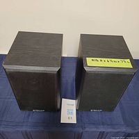 Front view of two black rectangular bookshelf speakers with wood-grain finish and black fabric grilles, placed on blue fabric surface with size note on one speaker.