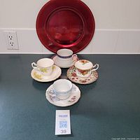 Four vintage teacups on matching saucers arranged in front of a red decorative charger plate, showing overall lot contents.