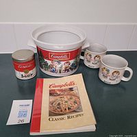 Photo of the lot on a countertop showing the soup tureen with winter children design, two matching mugs, a Campbell's Soup candle canister, and a classic recipe cookbook.