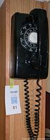 Black rotary dial wall phone mounted on wood panel with coiled cord and handset in place.