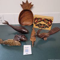 Lot of wooden art pieces on table, including various fish and bird carvings and a native art wooden panel