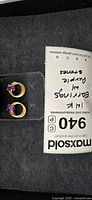 Pair of gold earrings with noted purple stones and multi-loop gold band design, placed on black surface with Maxsold tag.