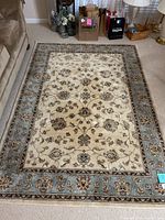 Full view of the rectangular area rug placed on the floor beside a sofa and some bags, showing the cream base with floral design and blue border.