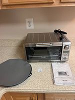 Stainless steel Breville toaster oven on a kitchen counter with a round baking pan and manuals beside it.