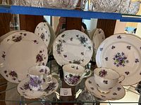 Set of china plates, teacups, and saucers with purple floral pattern and gold rims displayed on a glass shelf.