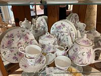 Full shot of the entire china tea set with teapot, sugar bowl, creamer, cups, saucers, and plates arranged on a glass shelf.