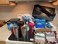 Photo of multiple items on stovetop including stainless steel insulated travel mugs, travel cups, ZeroWater water filter pitcher box, pink BlenderBottle shaker, replacement water filters and ice packs in a box.