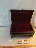 Jewelry box open showing velvet lining, earrings in various compartments, lucite necklace holder to left, small jewelry pieces inside box, bracelets and watch on table in front.