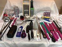 Photo showing a variety of kitchen utensils including ladles, graters, knives, vegetable peelers, measuring cup, and miscellaneous tools on a white backdrop.