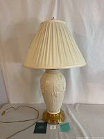 Full view of the Lenox table lamp showing the white ceramic base with floral embossing, brass-colored metal base, attached power cord, and cream pleated lampshade.