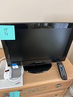 Front-facing view showing the entire Samsung 19-inch TV with attached base, remote control to the right, power cord, and user manual on the table underneath the TV screen.