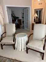 Two upholstered armchairs placed on either side of a round table with a cream fabric tablecloth and glass top in a living room.