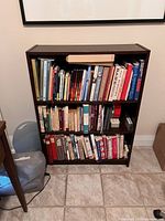 Front view of dark wood bookshelf with three shelves filled with books (not included in sale). Tiles floor background.