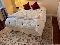Double mattress bed setup with embroidered white duvet, dark red pillows, beige heating blanket and bed skirt, placed on a floral area rug in a bedroom.