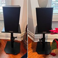 Pair of black Mission LX-1 MKII bookshelf speakers shown mounted on black adjustable metal stands. Speakers have black grilles with a rectangular form. Stands are height adjustable with locking knobs and hexagonal bases for stability.