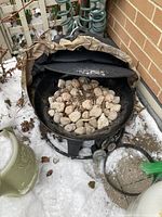 Single photo of a black propane outdoor firepit placed on snowy ground near a brick wall. The firepit is filled with lava rocks and has a brown cover partially pulled back. Shows signs of outdoor use and storage.