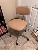 Photo showing front and left side view of vintage beige fabric office chair on caster wheels, positioned near bookshelf and door.